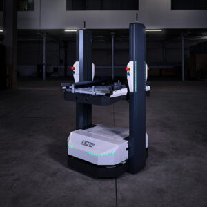 Open Shuttle 100 – AMR para el transporte de cajas En un espacio industrial con poca luz se encuentra un vehículo robótico de transporte para almacenes, provisto de columnas verticales y cintas transportadoras.