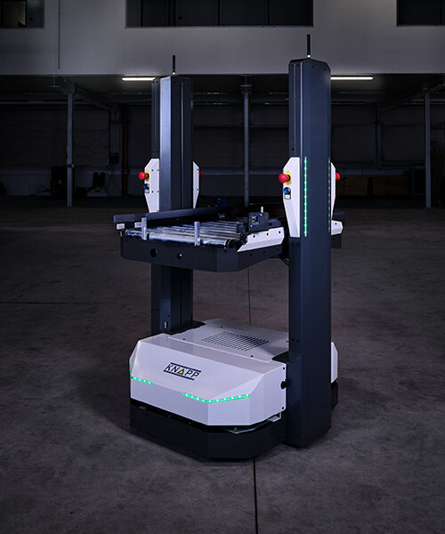 Open Shuttle 100 – AMR para el transporte de cajas En un espacio industrial con poca luz se encuentra un vehículo robótico de transporte para almacenes, provisto de columnas verticales y cintas transportadoras.