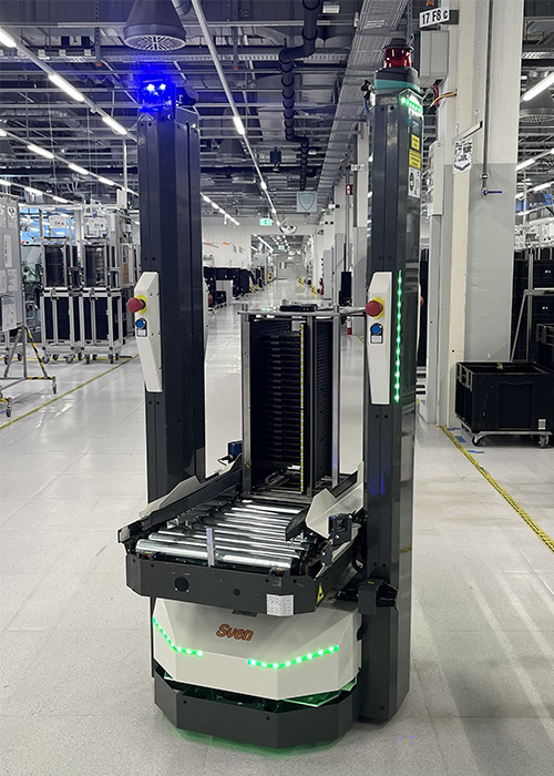 Open Shuttle ASG scaling Autonomous robot with conveyor platform and green lights operates in a modern industrial facility with machinery and bright overhead lighting.