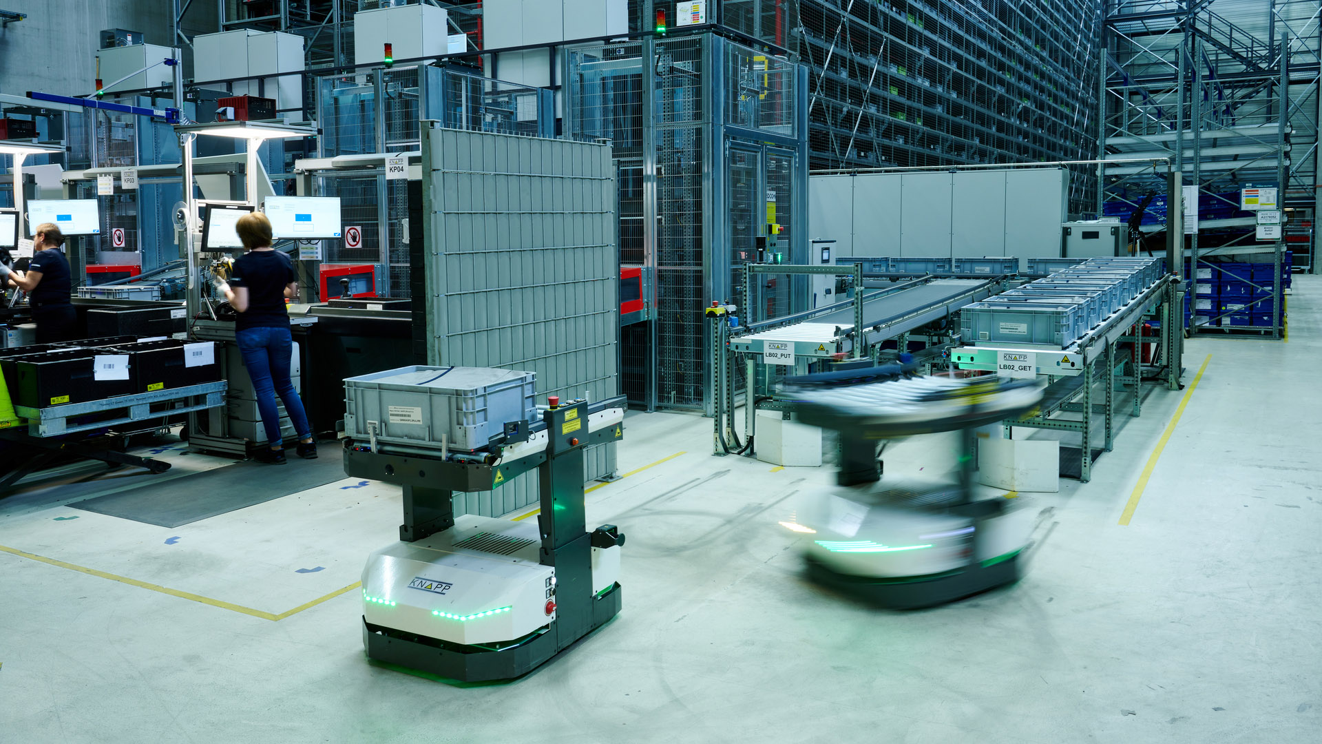Zwei Roboter transportieren Behälter in einem modernen Lager, während im Hintergrund Mitarbeiter an Computerarbeitsplätzen sitzen und Regale mit Behältern gefüllt sind.
