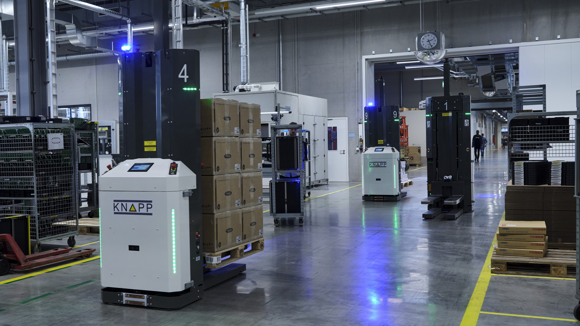 Open Shuttle Fork Flotte Warehouse robots transport stacked boxes on pallets in a large, brightly lit facility with shelves and equipment in the background.