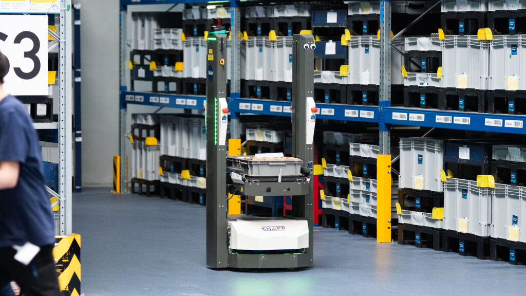 Ein mobiler Roboter transportiert Artikel durch einen Lagergang und verdeutlicht die Vorteile und Kosten eines fahrerlosen Transportsystems.