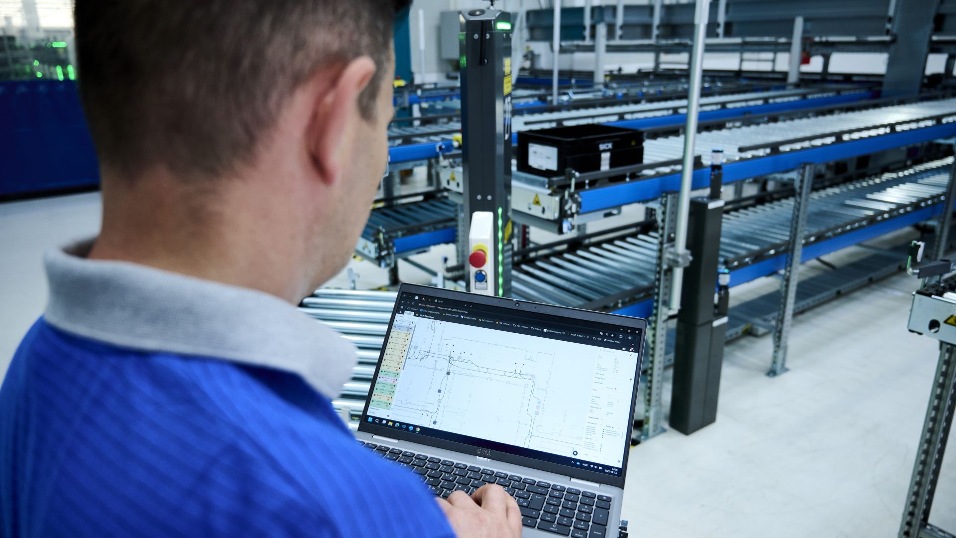 Eine Person betrachtet auf einem Laptop ein Layout einer Anlage, in der im Hintergrund Förderbänder und autonome mobile Roboter im Einsatz sind.