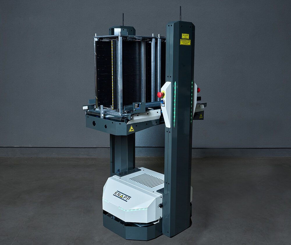 Un robot industrial alto, con una base blanca y una columna vertical, que se utiliza como AMR para el transporte de portaplacas, se encuentra frente a una pared gris lisa.