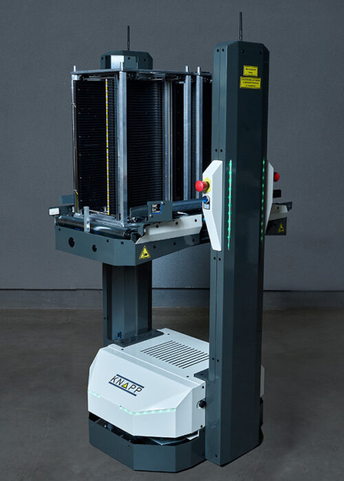 Open Shuttle ASG for magazine transport Robot industrial alto y gris, con una base blanca y una estructura vertical negra, situado sobre un suelo de hormigón frente a una pared gris lisa.