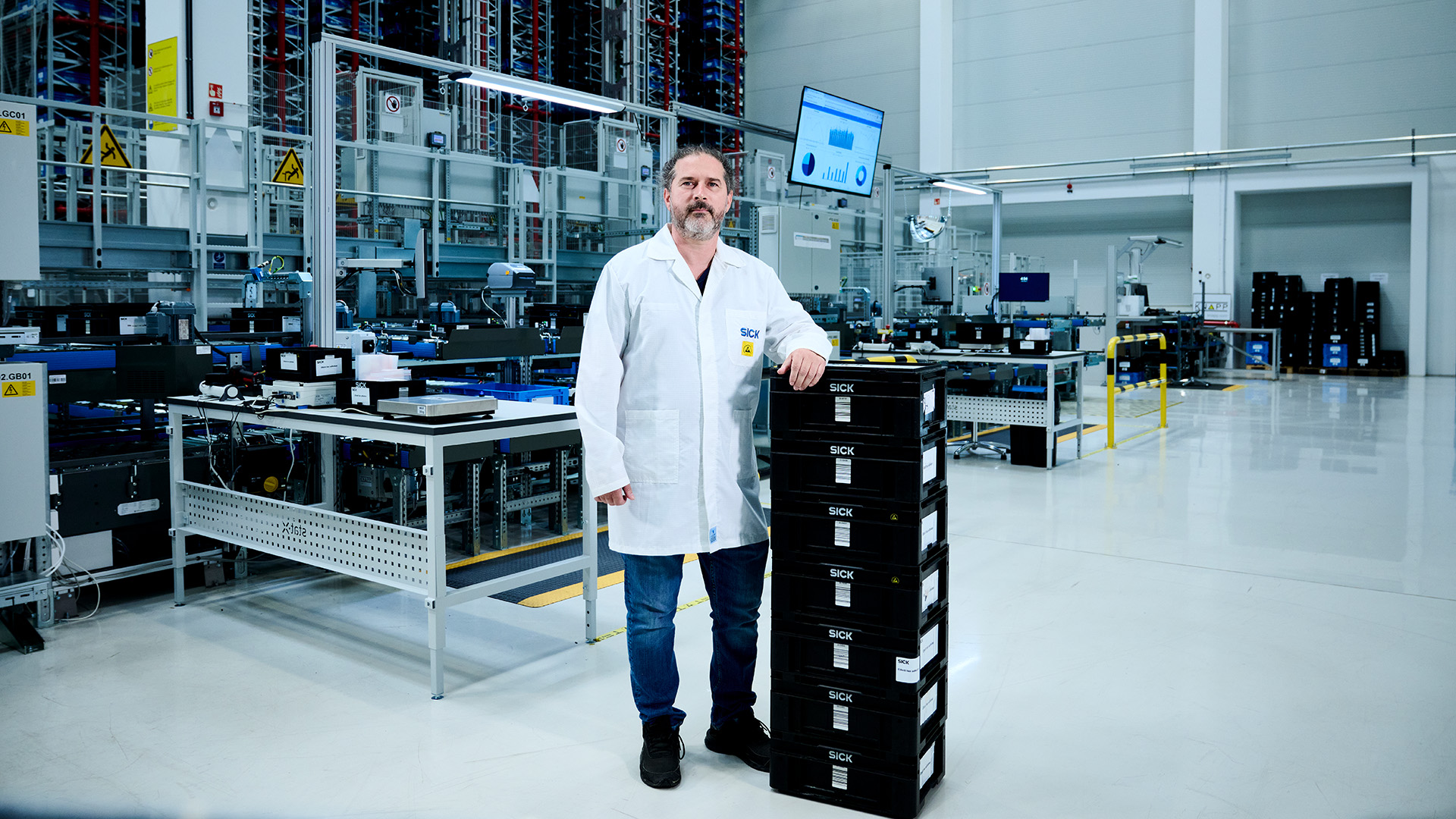 Un homme en blouse blanche se tient à côté d'une pile de conteneurs noirs dans un entrepôt industriel moderne de haute technologie, avec des machines, des équipements et un moniteur visibles à l'arrière-plan.