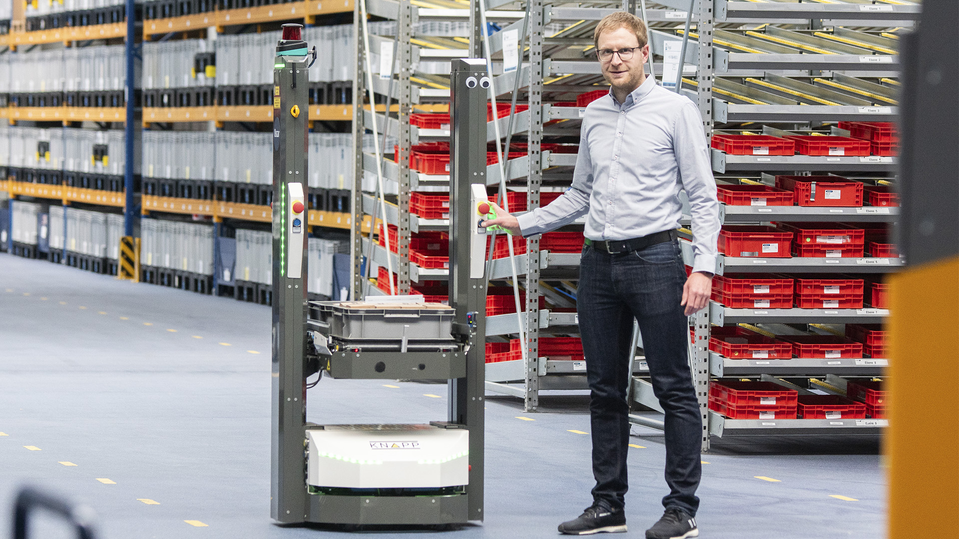 Pascal Weiland at Schunk with the Open Shuttle A man with strawberry blond hair and glasses stands next a KNAPP mobile robot carrying containers.