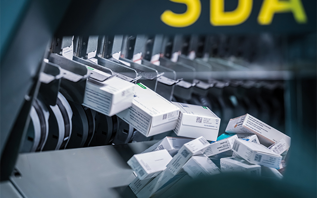 Peak_pharmacy_lager_SDA_medikamente_close_up Plusieurs boîtes de médicaments sont distribuées par une machine automatisée et tombent dans une zone de collecte située en contrebas. La scène suggère un processus organisé et mécanisé de tri ou de distribution de médicaments.