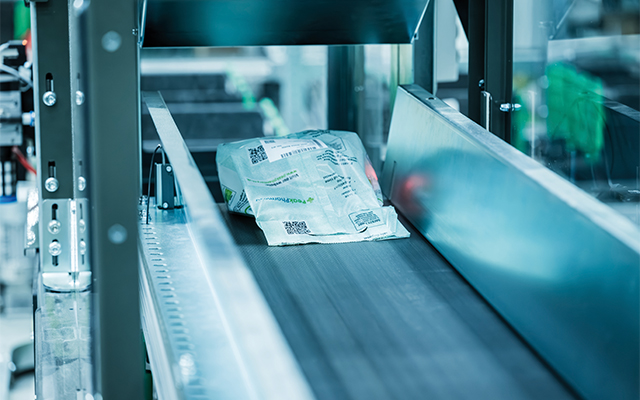 Peak_pharmacy_medikamenten_transport Une enveloppe matelassée avec des codes-barres et des étiquettes imprimés se déplace sur un tapis roulant dans une installation d'emballage ou de triage automatisée.