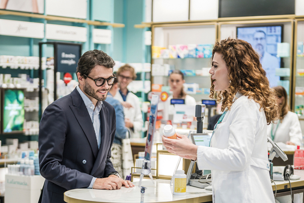 A pharmacist in a white coat explains a product to a smiling customer at a pharmacy counter, with shelves of medicine in the background.