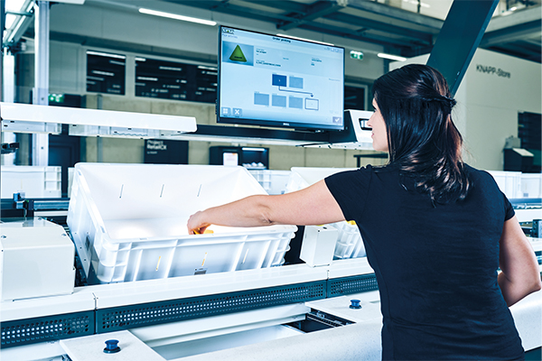pick-it-easy-workstations-in-action A woman in a black shirt inspects yellow produce on a conveyor belt in a modern, automated factory setting with machinery in the background.