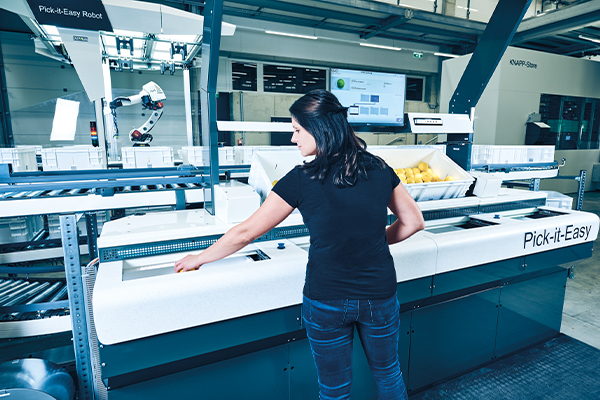 Eine Frau bedient eine automatische Sortiermaschine, wobei sie die Anweisungen auf einem Computermonitor über den Lagerbehältern in einer Industrieumgebung sieht.