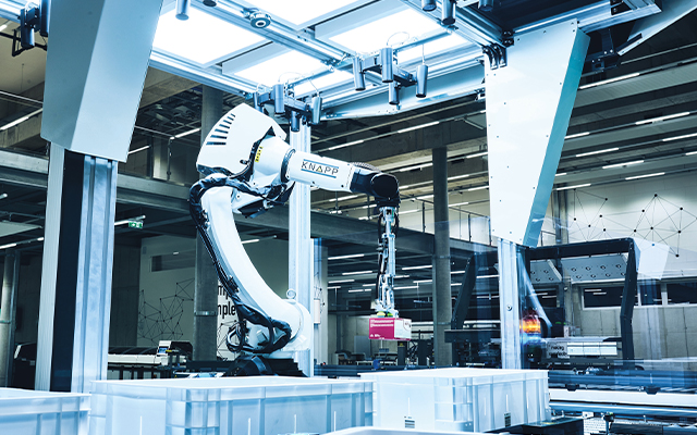 pick_it_easy_roboter Dans une installation industrielle moderne, un bras robotisé déplace une petite boîte sur un conteneur, entouré de parois en verre, de poutres métalliques et d'un éclairage zénithal intense.