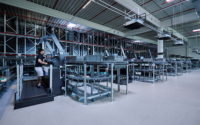pick_it_easy_workstations Une personne manipule un panneau de commande à côté de bandes transporteuses automatisées et de rayonnages de stockage dans un entrepôt moderne et spacieux avec de hauts plafonds et un éclairage industriel.