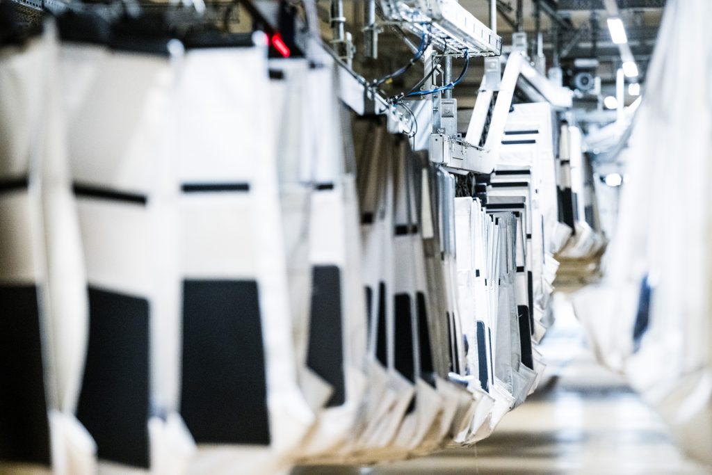 Des rangées de grands sacs en tissu ou de sacs à linge sont suspendues à un système de convoyage automatisé dans une installation industrielle, avec un éclairage brillant et des structures métalliques visibles à l'arrière-plan.