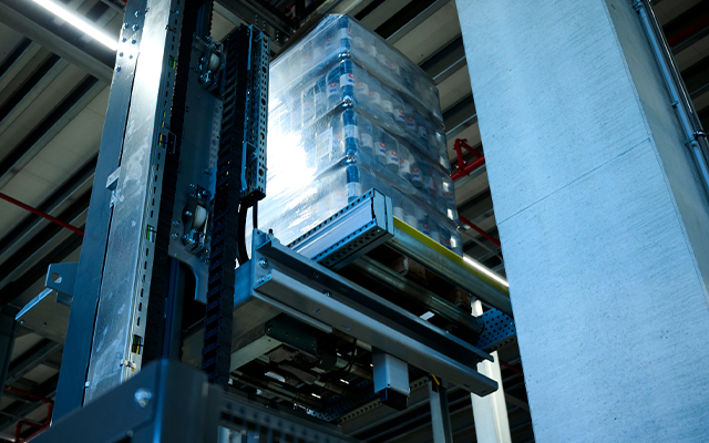 Powerline_beitragsbild A robotic lift raises a pallet stacked with shrink-wrapped goods in a modern warehouse, surrounded by metal beams and concrete columns.