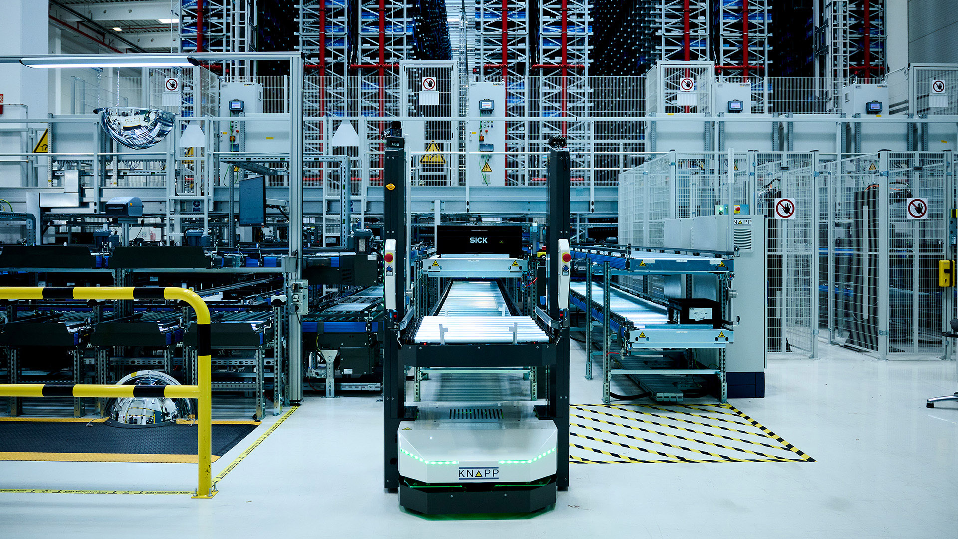 Automatisierte Roboter und Förderbänder ergänzen die SICK Produktionslogistik in einem modernen Hightech-Lager mit Regalen und Maschinen im Hintergrund.