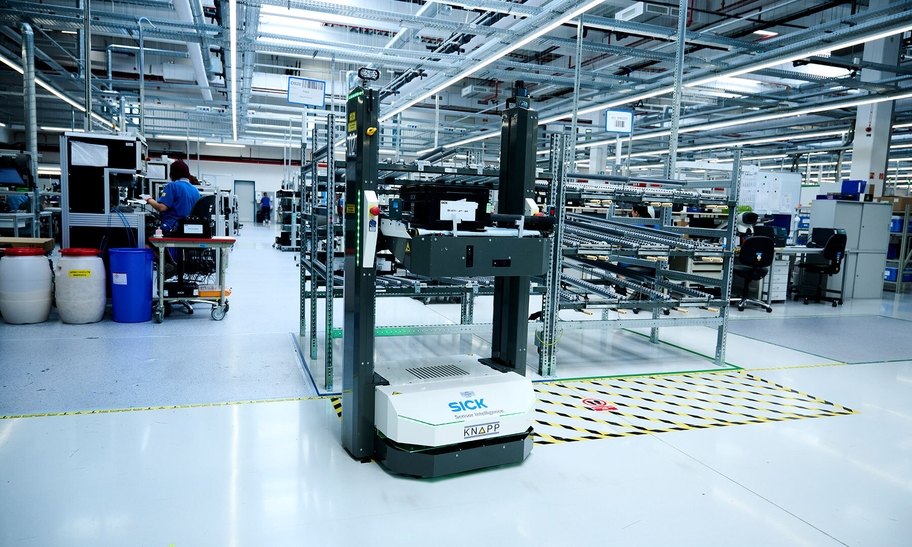 Un véhicule à guidage automatique (AGV) appelé "SICK" navigue dans une usine spacieuse et moderne avec des équipements industriels, des postes de travail et un ouvrier visible à l'arrière-plan.