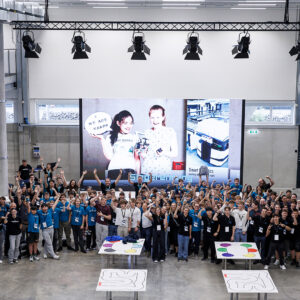 Eine große Gruppe jubelt bei einer Indoor-Veranstaltung zum Thema Automatisierungslösungen für die gesamte Wertschöpfungskette, mit Robotikmatten und -geräten auf dem Boden.