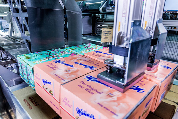 Runpick_beitragsbild Ein Palettierroboter Runpick verpackt effizient Stapel von bunten Kartons mit der Aufschrift "Waiwera" in einer automatisierten Produktionsanlage und drückt sie nach unten, während sie sich auf dem Fließband bewegen.