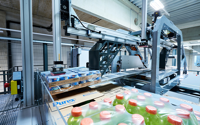 Runpick_beitragsbild_02 A modern automated packaging machine and a Palletizing robot Runpick wrap pallets of bottled drinks and boxes inside an industrial warehouse setting with metal beams and bright lighting.