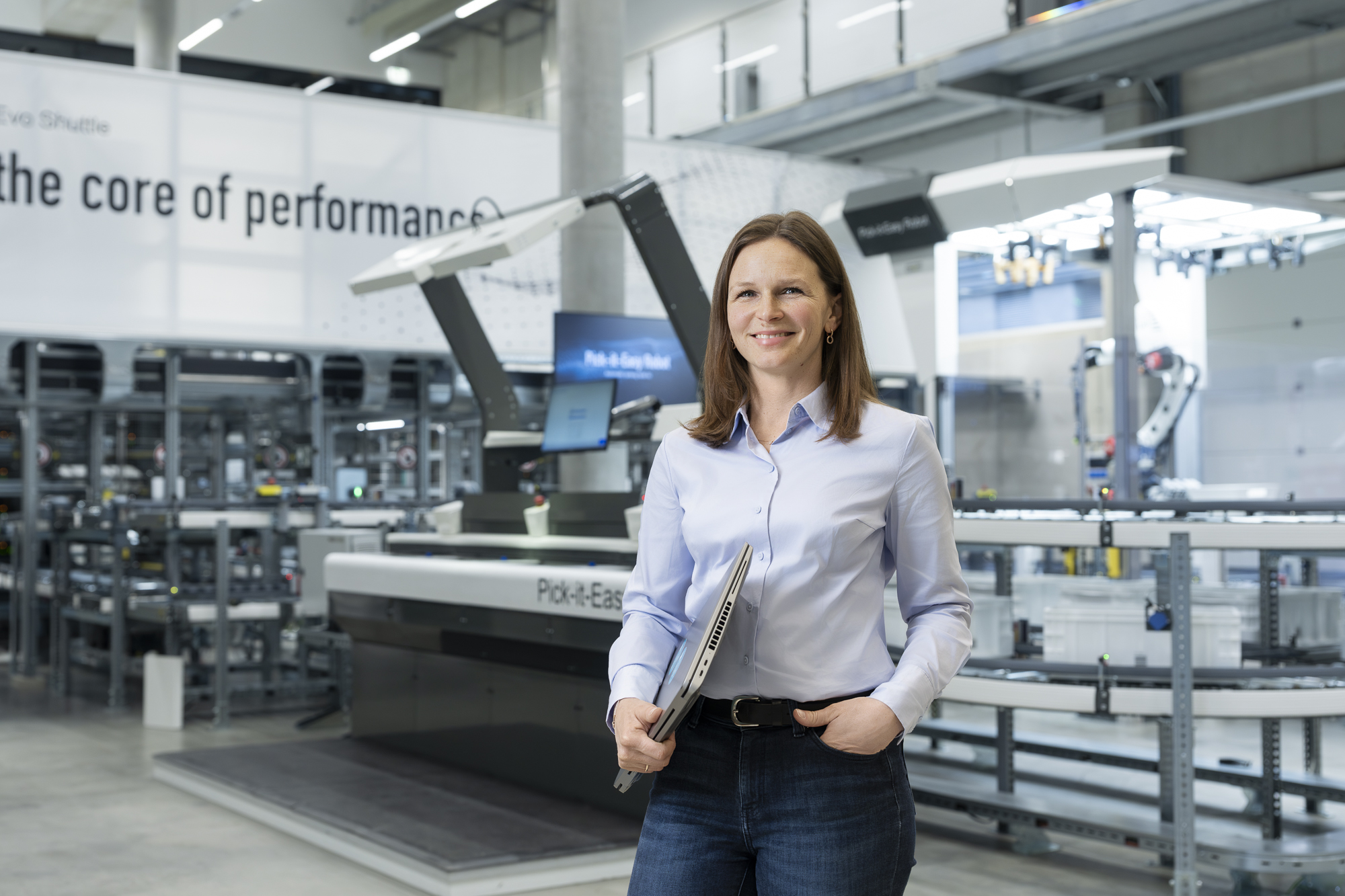 Une femme en chemise bleue se tient dans une installation industrielle moderne, tenant un ordinateur portable et souriant, avec des machines et des équipements en arrière-plan.