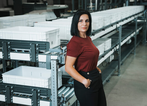 A woman with dark hair stands confidently with her hands in her pockets in front of a conveyor belt system with white plastic bins in a factory or warehouse setting.