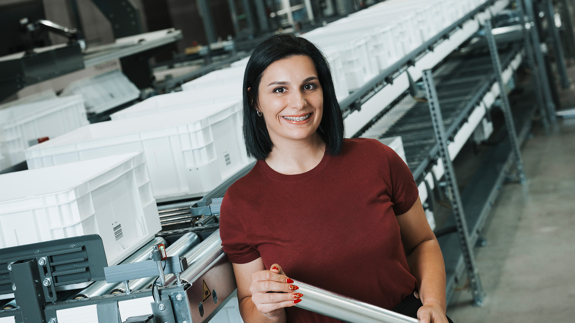 A woman with short dark hair, wearing a maroon t-shirt, stands smiling in a factory or warehouse setting, surrounded by white bins and metal conveyor belts.