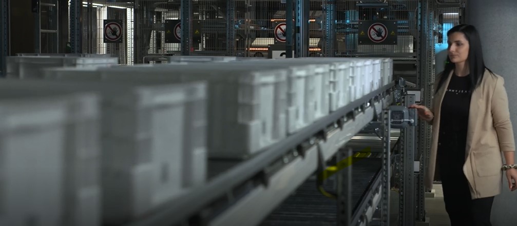 A woman stands beside a conveyor system with multiple white storage bins in an industrial or warehouse setting. No photography signs are visible on the wall in the background.
