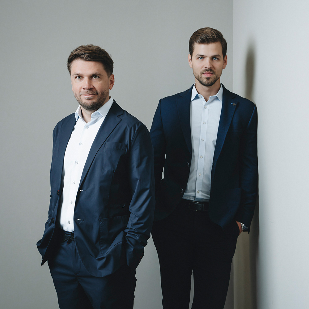 Sascha_Seruga_und_Thomas_Zilk Zwei Männer in dunklen Anzügen und weißen Hemden stehen in einem Innenraum vor einer schlichten hellen Wand. Als Partner und Integratoren für Lagerlogistik blicken sie mit leichtem Lächeln in die Kamera - der eine mit den Händen in den Taschen, der andere steht direkt hinter ihm, die Arme entspannt.