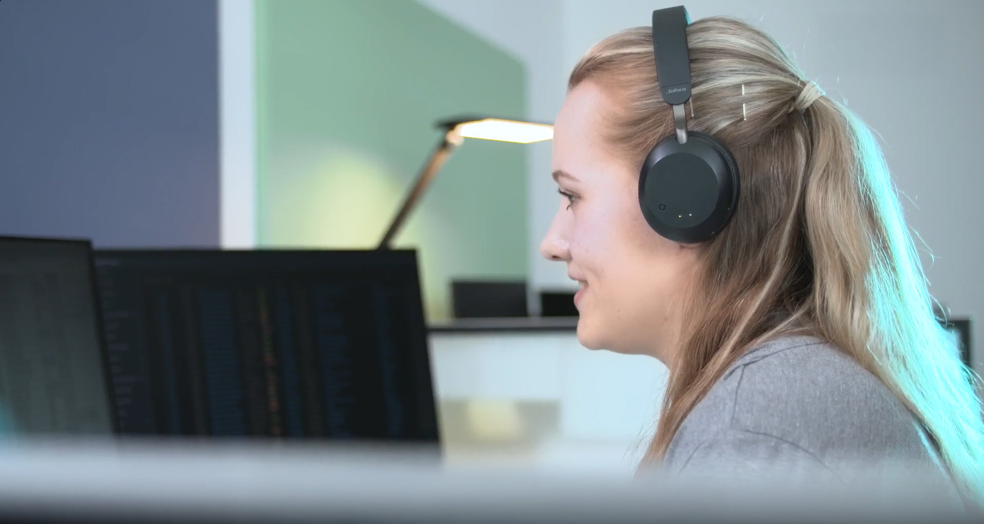 Our colleague Saskia has headphones on and is smiling as she looks at a computer screen in an office environment. A desk lamp and another monitor can be seen in the background.