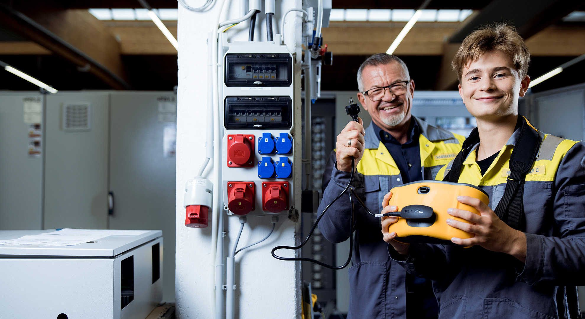 An apprentice and trainer hold a measuring device and a cable and smile at the camera.