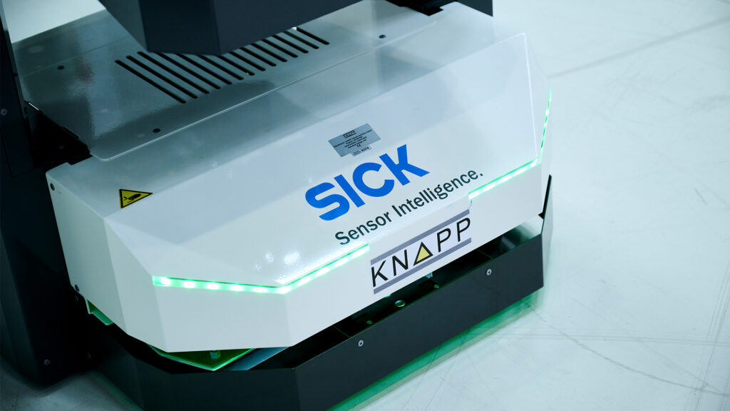 Un appareil industriel blanc portant les logos KNAPP et SICK Sensor Intelligence, avec des diodes électroluminescentes vertes à sa base, repose sur un sol propre et clair.