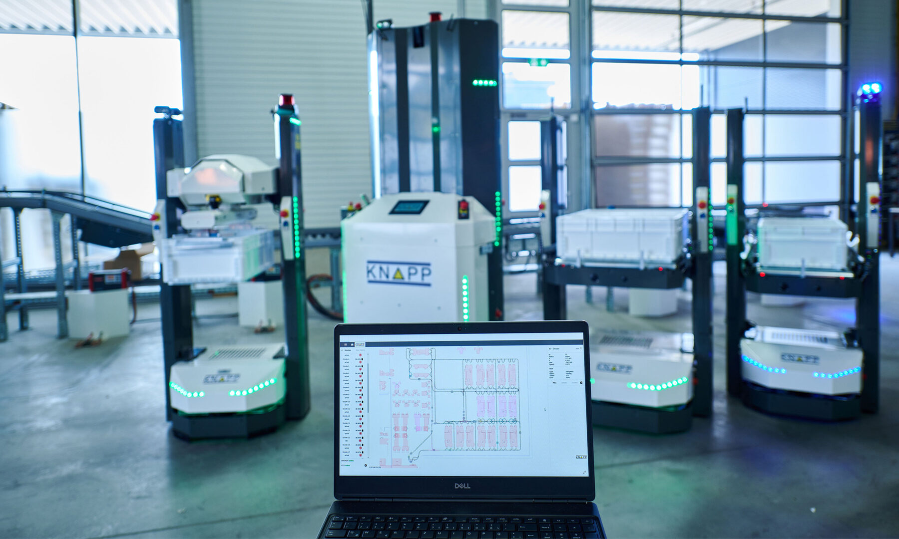 Software para AMR KiSoft FCS Un ordenador portátil muestra planos del almacén frente a cuatro robots automatizados con contenedores en el interior de unas modernas instalaciones industriales.