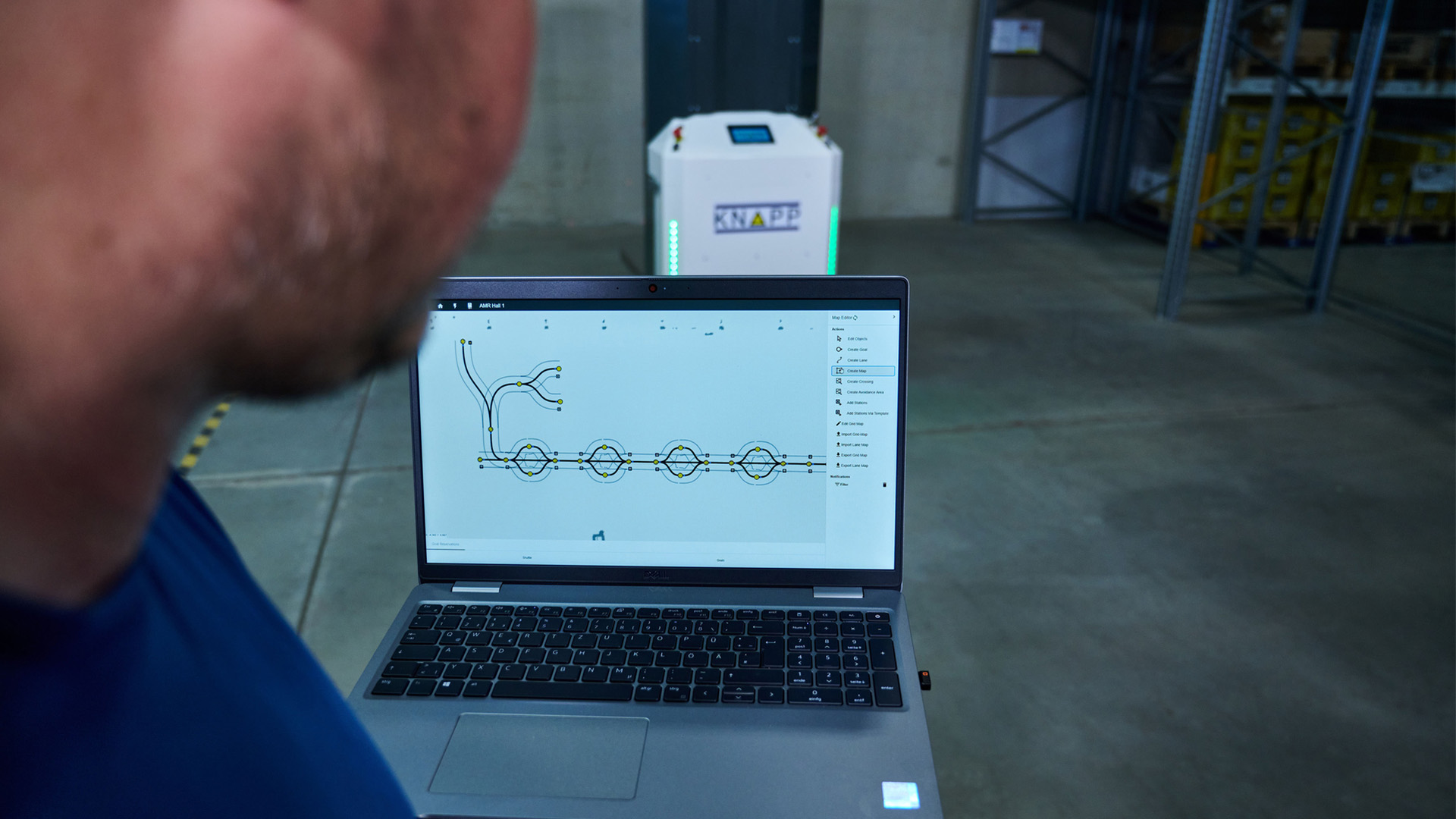 KiSoft FCS software for AMR pallet transport A person views a laptop displaying a logistics or warehouse layout, with a robot and shelves visible in the background.