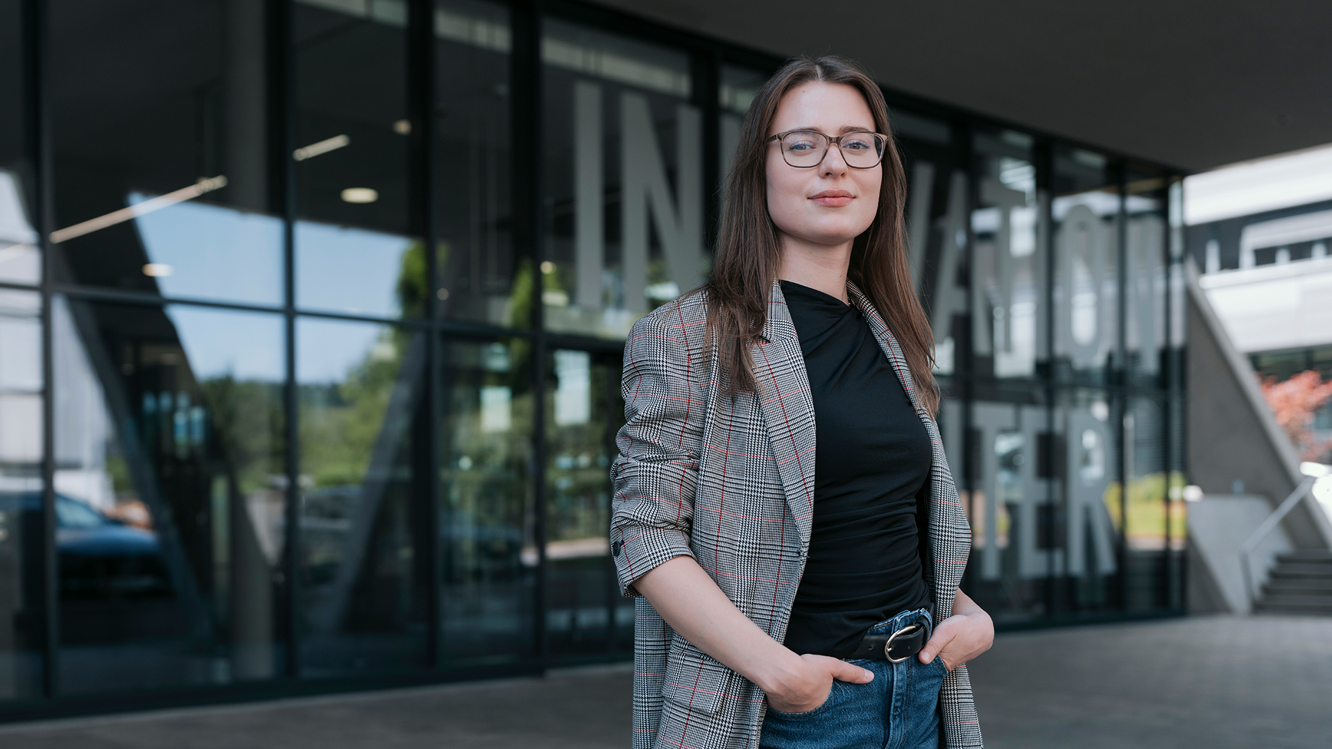 Eine Frau mit langen braunen Haaren und Brille steht selbstbewusst vor einem modernen Glasgebäude.