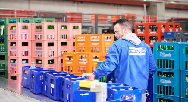 Front view of a sorting lane. The empty handling units are conveyed to the sorting lanes, where an employee takes the reusables from the conveyor and stacks them on unmixed pallets behind them. This image shows several beverage crates arriving at the work station, at a perfect height for the male employee to pick up.