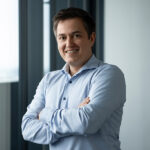 Ein lächelnder Mann in einem hellblauen Hemd steht mit verschränkten Armen in einem Büro, das auf Fahrerlose Transportsysteme spezialisiert ist.