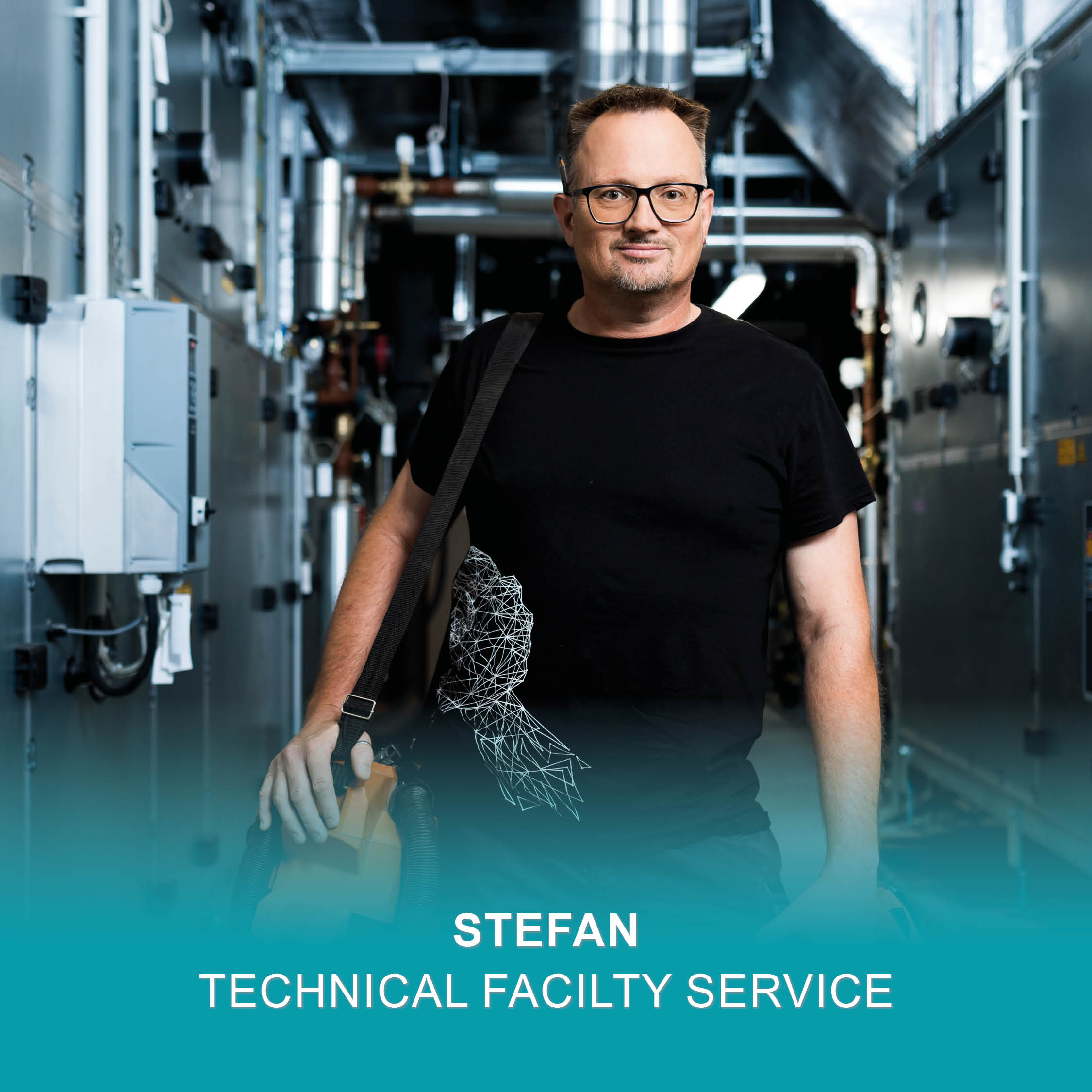 Un homme portant des lunettes et un t-shirt noir se tient dans un couloir d'une installation industrielle, transportant du matériel.