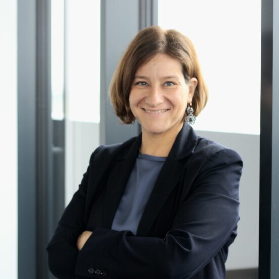 Eine Frau mit kurzen braunen Haaren, die einen dunklen Blazer und ein blaues Hemd trägt, steht in einem Raum, hat die Arme verschränkt und lächelt in die Kamera.