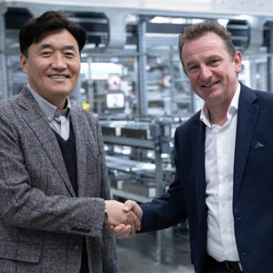 Zwei Männer in Anzügen schütteln sich in einer Fabrik die Hände und besiegeln inmitten von Metallgeräten einen Vertrag über Automatisierungslösungen für die gesamte Wertschöpfungskette.