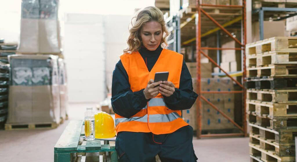 Woman is sitting in a warehouse and using the redPILOT teamAPP on her smartphone, an app for shift scheduling in the Operational Excellence Software.