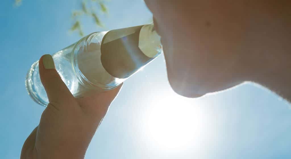 The image shows a woman in sunshine drinking a beverage from a glass bottle. The sky is deep blue. The glass bottle is in focus.