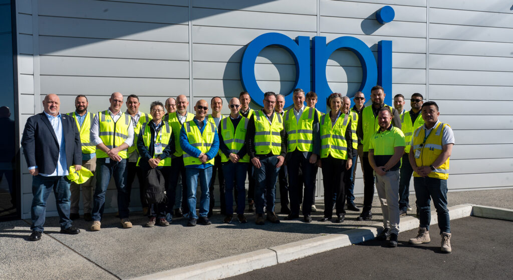 Un groupe de personnes portant des gilets de sécurité jaunes se tiennent ensemble à l'extérieur d'un bâtiment moderne avec les grandes lettres bleues "api" sur le mur derrière eux. Le groupe pose pour la photo par une journée ensoleillée.