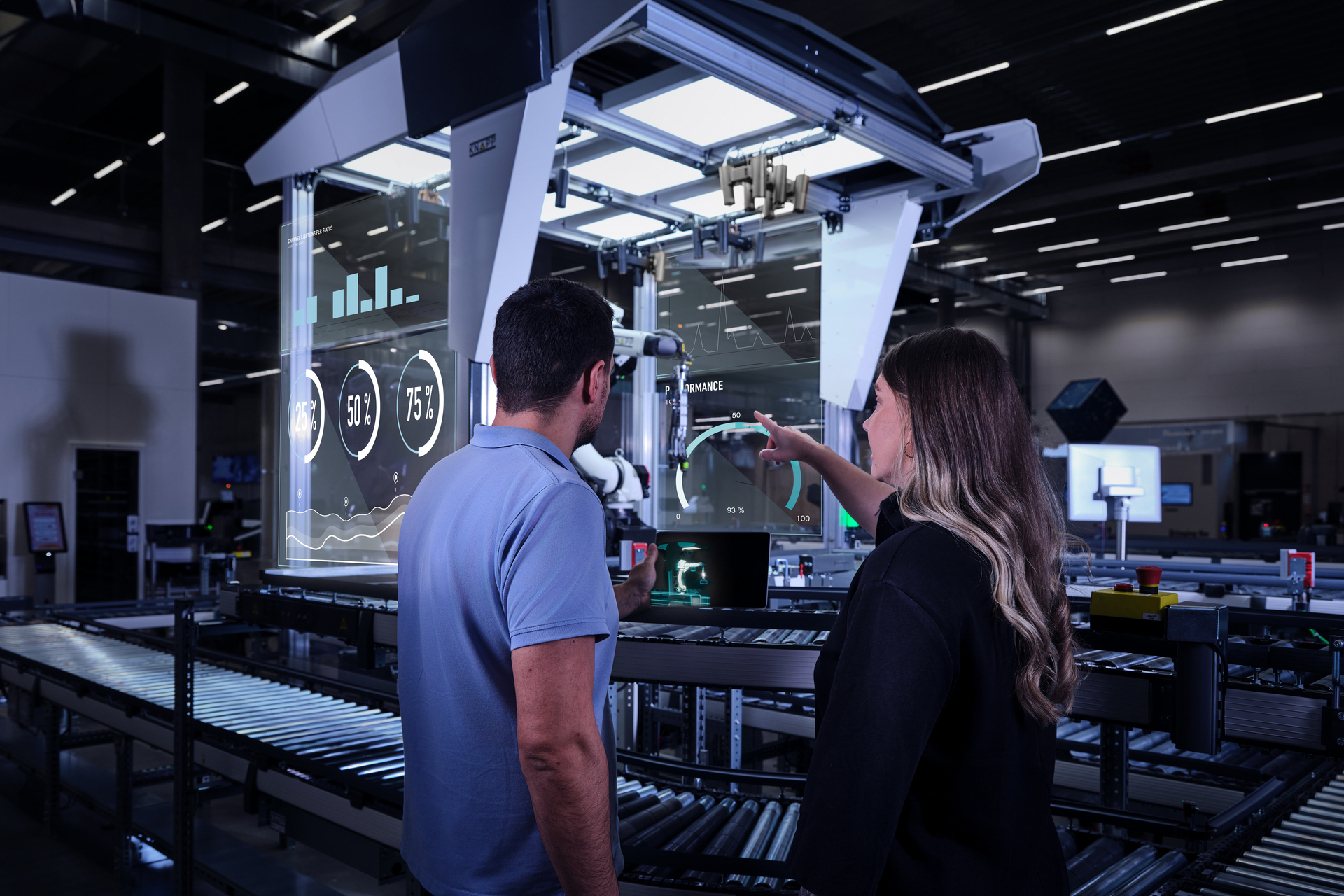 Plattformen_Software_hero Zwei Personen stehen an einem automatisierten Arbeitsplatz mit einem Roboterarm und großen transparenten Bildschirmen, auf denen Diagramme und Daten aus einer Lieferkettensoftware in einer modernen Fabrikumgebung angezeigt werden. Eine Person deutet auf den Bildschirm.