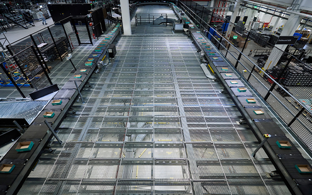 verpackungsgroessen Un grand système de tri automatisé dans un entrepôt, avec des tapis roulants transportant de petits colis, entouré de rampes métalliques et d'équipements industriels dans une installation spacieuse et bien éclairée.