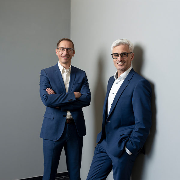 Deux hommes en costume bleu, représentant Logistique Fashion, se tiennent contre un mur gris clair, l'un les bras croisés, l'autre les mains dans les poches.