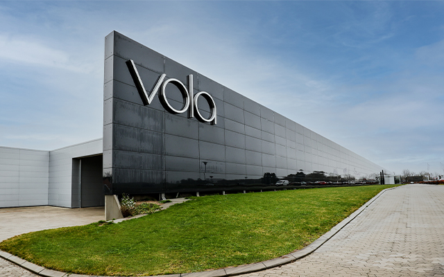 A modern, black-paneled building with the large white letters “Vola” on its side, set against a partly cloudy sky with a green lawn and paved driveway in the foreground.