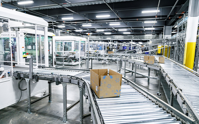 Une boîte en carton se déplace sur un tapis roulant métallique dans un entrepôt ou un centre de distribution moderne et bien éclairé, avec des machines automatisées en arrière-plan.