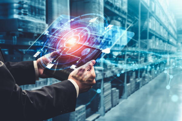 Eine Person im Anzug verwendet ein digitales Tablet mit einer holografischen Schnittstelle, um in einem modernen Lagerhaus das Inventar mit einer Lieferkettensoftware zu verwalten.